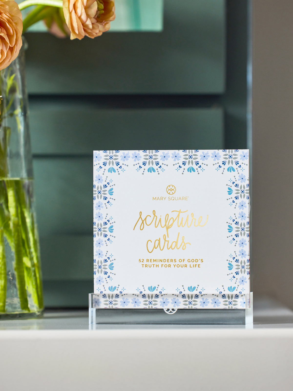 A boxed set of Mary Square scripture cards displayed on a clear acrylic stand on a shelf, with a blurred teal cabinet in the background and peach flowers in a glass vase at the left edge.
