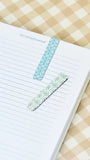 Two decorative washi tape bookmarks with scalloped edges, one light blue and one white with tiny multicolor floral prints, shown on a lined journal page over a beige and white gingham fabric background.