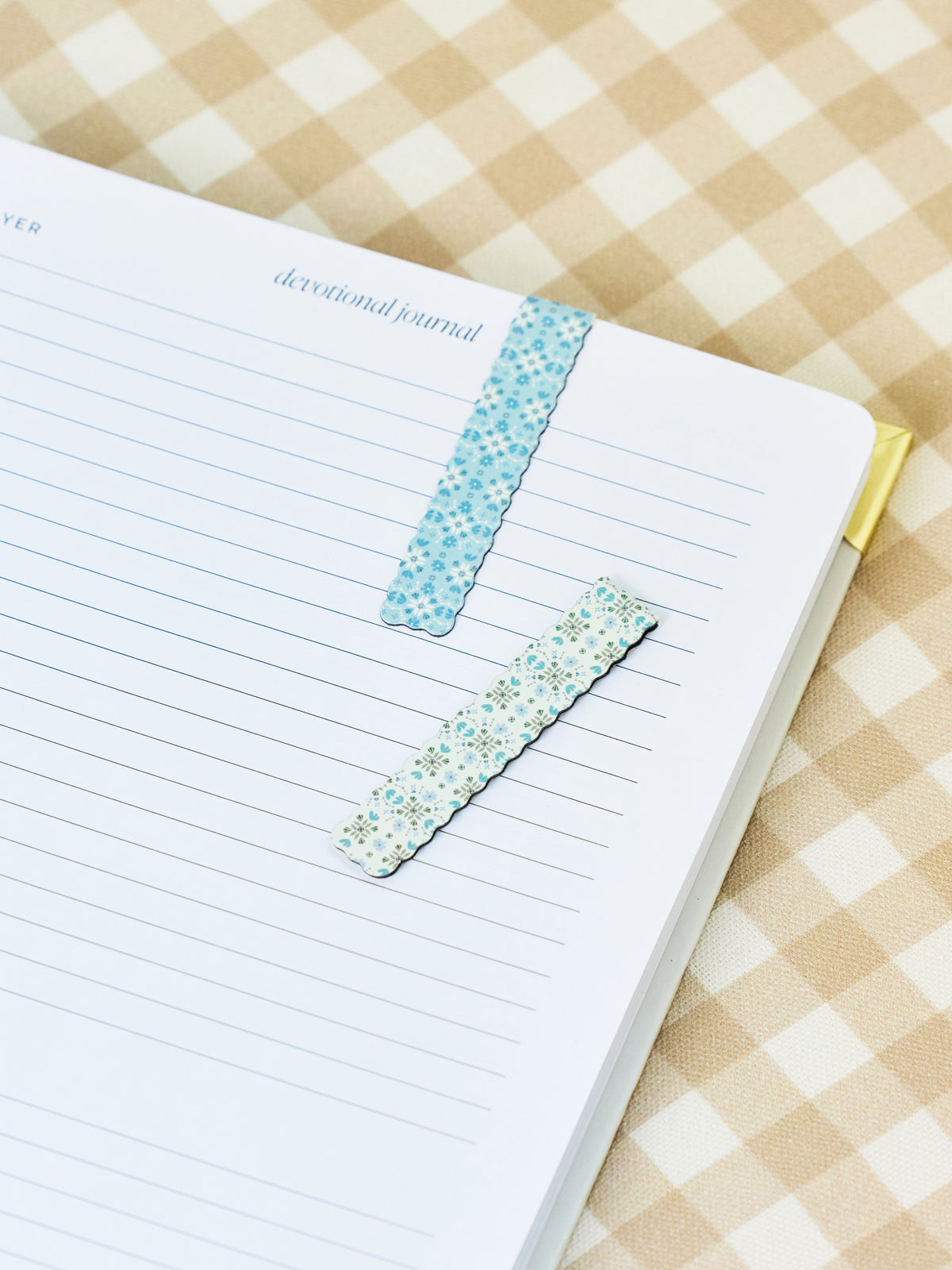 Two decorative washi tape bookmarks with scalloped edges, one light blue and one white with tiny multicolor floral prints, shown on a lined journal page over a beige and white gingham fabric background.