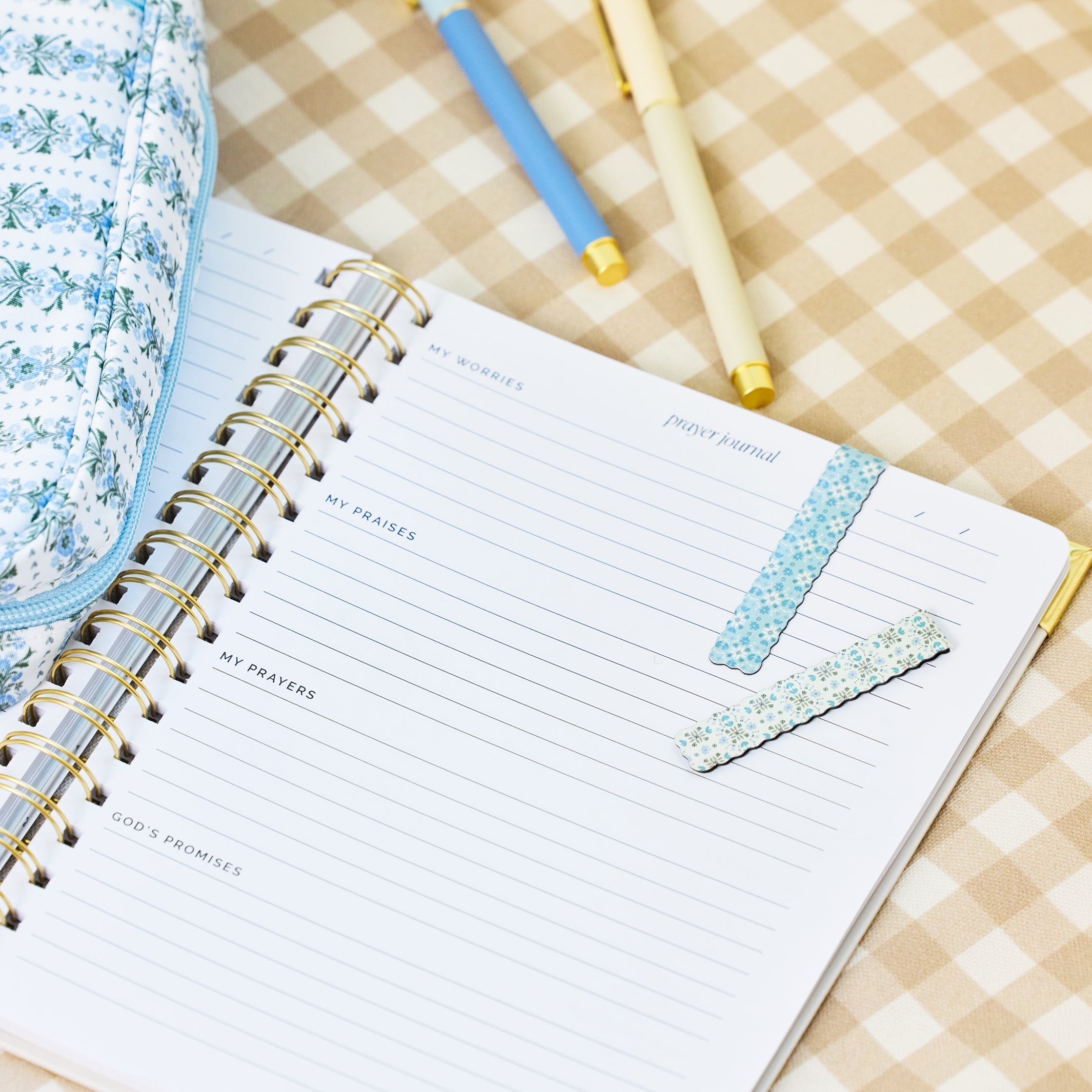 An open spiral-bound prayer journal with lined pages lies on a beige-and-white gingham surface, styled with two pastel pens and a matching patterned pencil case.