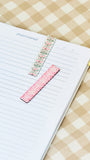 Two decorative floral washi tape bookmarks, one pink and one green, placed on an open lined prayer journal on a beige and white checkered fabric background.
