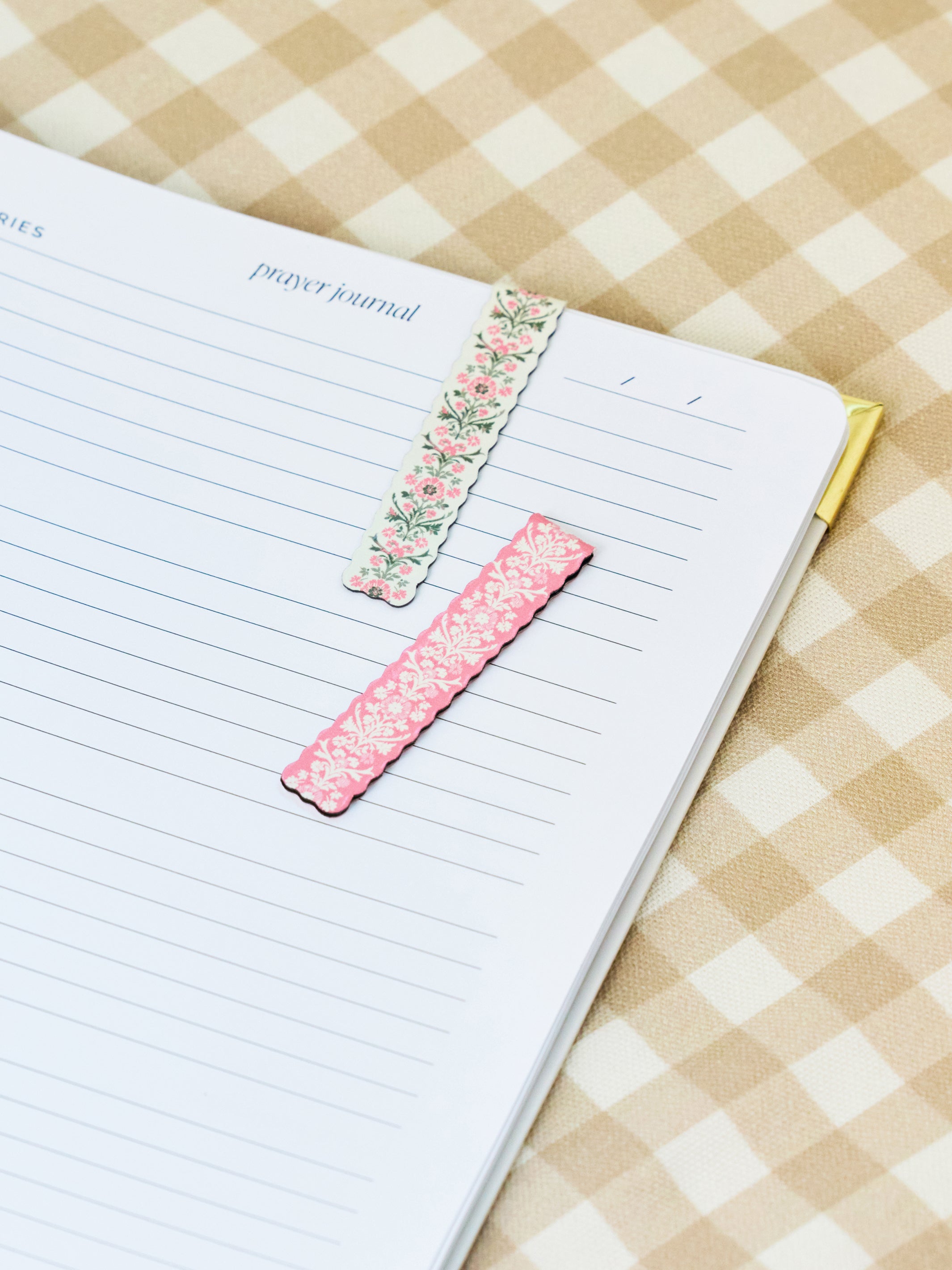 Two decorative floral washi tape bookmarks, one pink and one green, placed on an open lined prayer journal on a beige and white checkered fabric background.