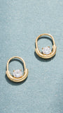 A pair of gold hoop-style earrings with a central oval setting and a clear faceted gemstone, displayed on a textured light blue surface.