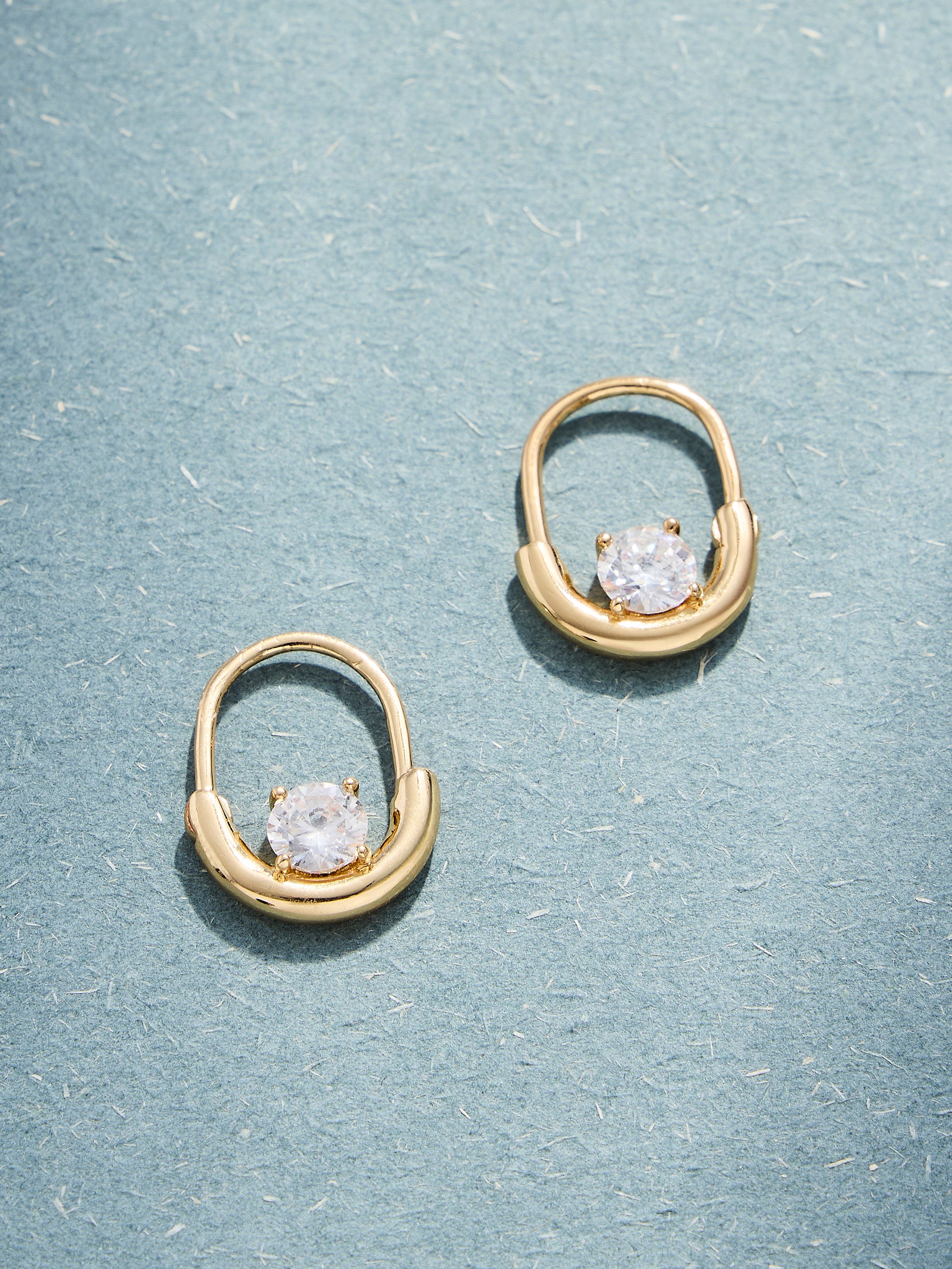 A pair of gold hoop-style earrings with a central oval setting and a clear faceted gemstone, displayed on a textured light blue surface.
