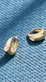 A pair of gold huggie hoop earrings with teardrop-shaped clear crystals, displayed on a textured blue woven surface.