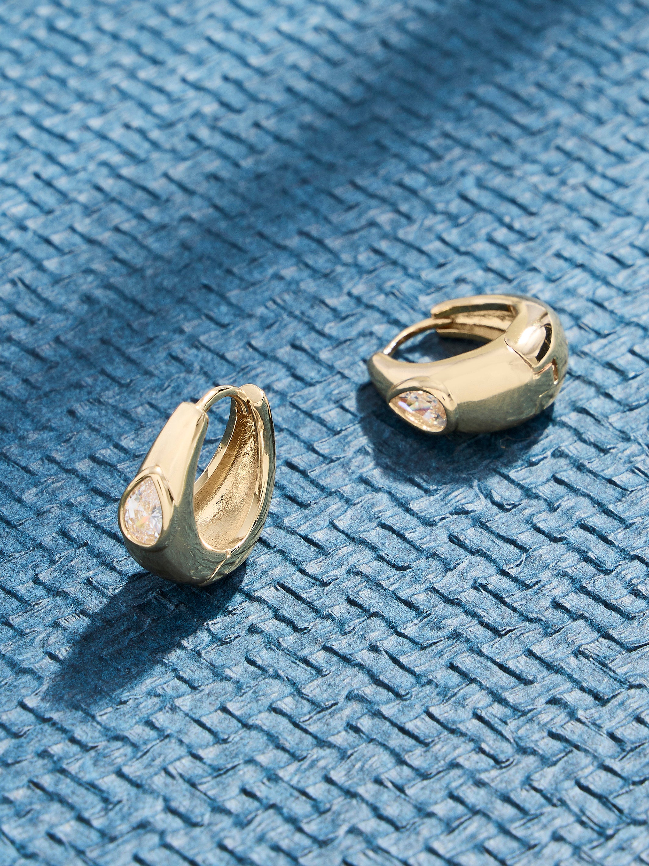 A pair of gold huggie hoop earrings with teardrop-shaped clear crystals, displayed on a textured blue woven surface.