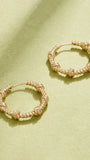 A pair of gold-toned textured hoop earrings with knotted details, displayed on a textured light green surface with soft shadows.