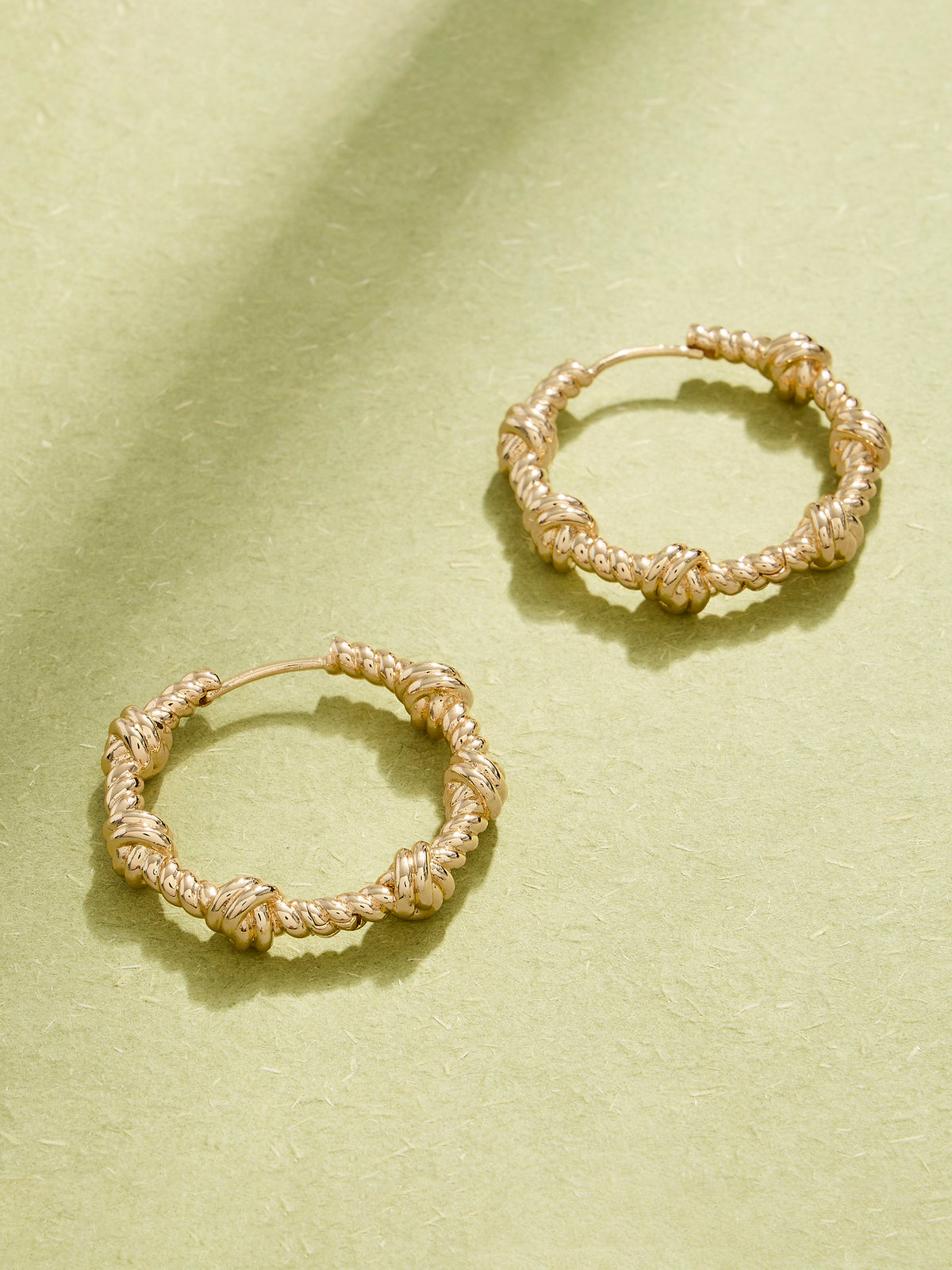 A pair of gold-toned textured hoop earrings with knotted details, displayed on a textured light green surface with soft shadows.