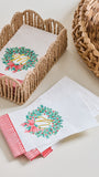 White paper napkins with a green wreath and red bow design, arranged in woven baskets on a light beige surface.