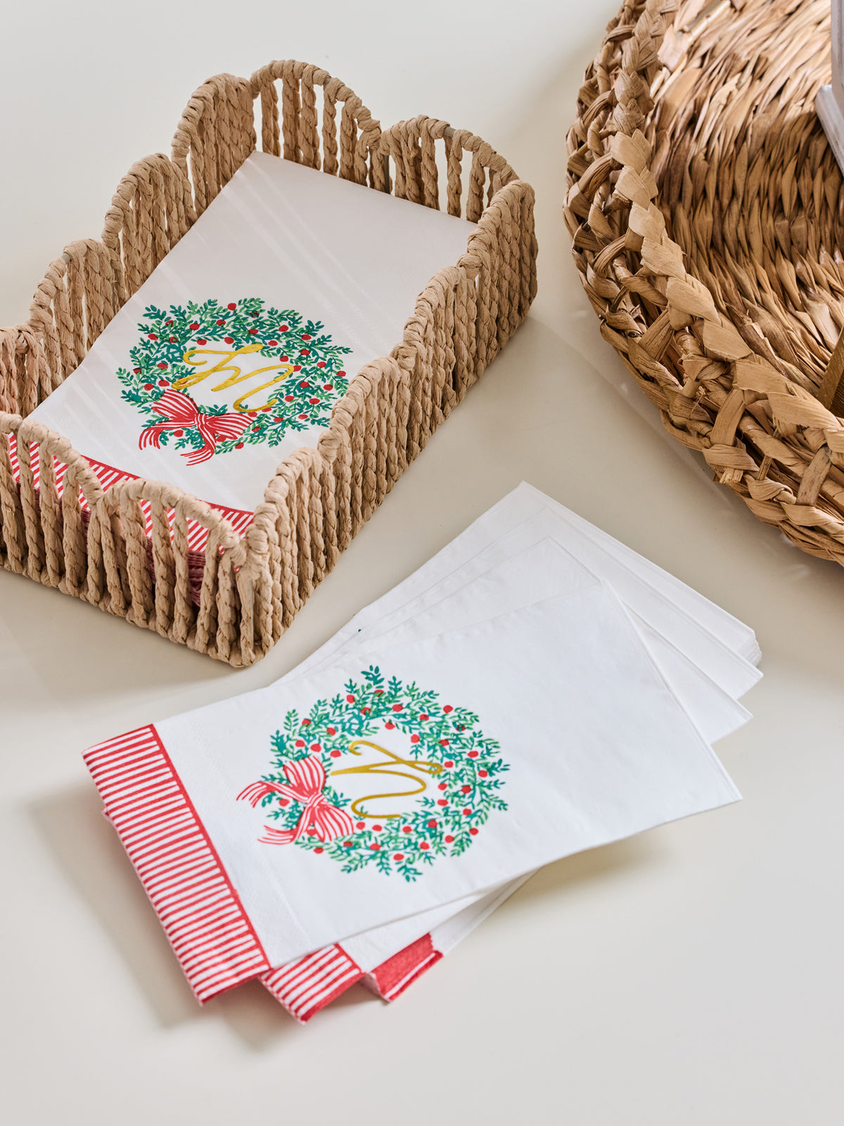 White paper napkins with a green wreath and red bow design, arranged in woven baskets on a light beige surface.