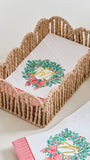 A woven rope baby carrier basket holding a white blanket with a green holly wreath and red bow design, with a matching folded blanket and striped fabric visible in the foreground on a light neutral background.