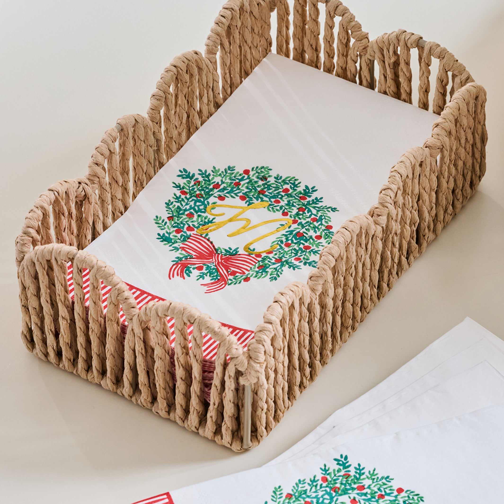A woven rope baby carrier basket holding a white blanket with a green holly wreath and red bow design, with a matching folded blanket and striped fabric visible in the foreground on a light neutral background.