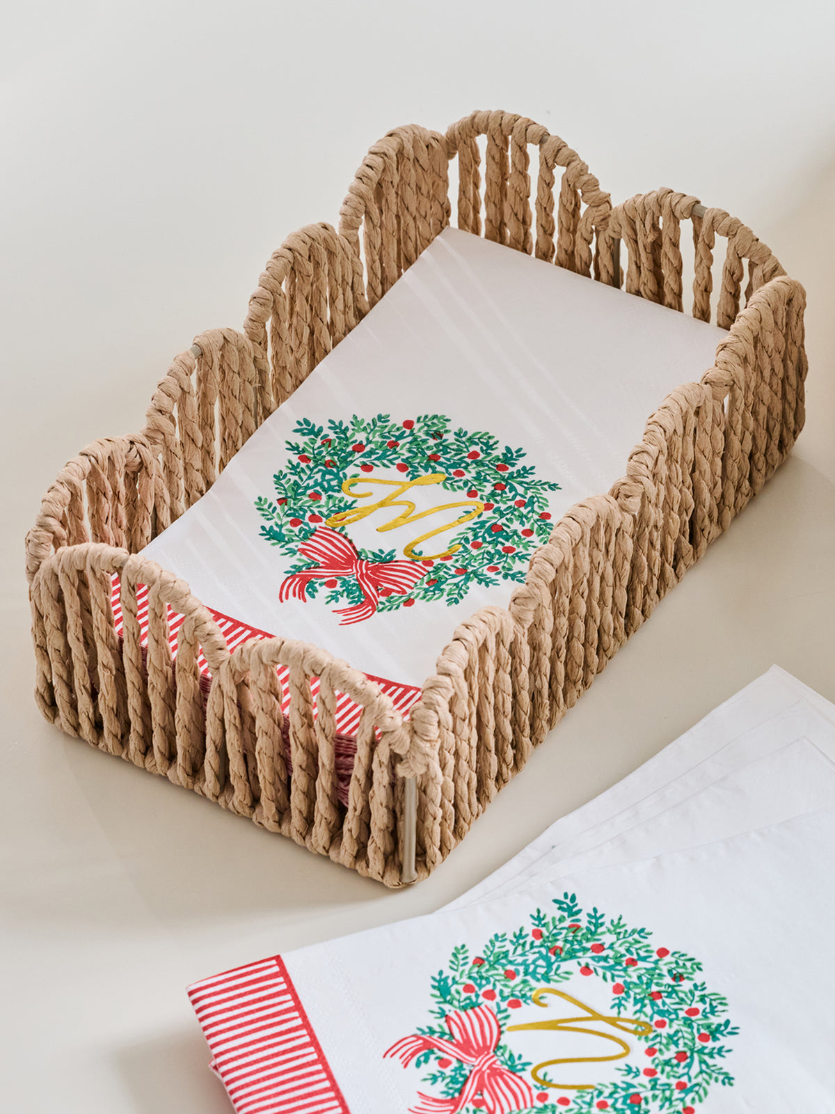A woven rope baby carrier basket holding a white blanket with a green holly wreath and red bow design, with a matching folded blanket and striped fabric visible in the foreground on a light neutral background.