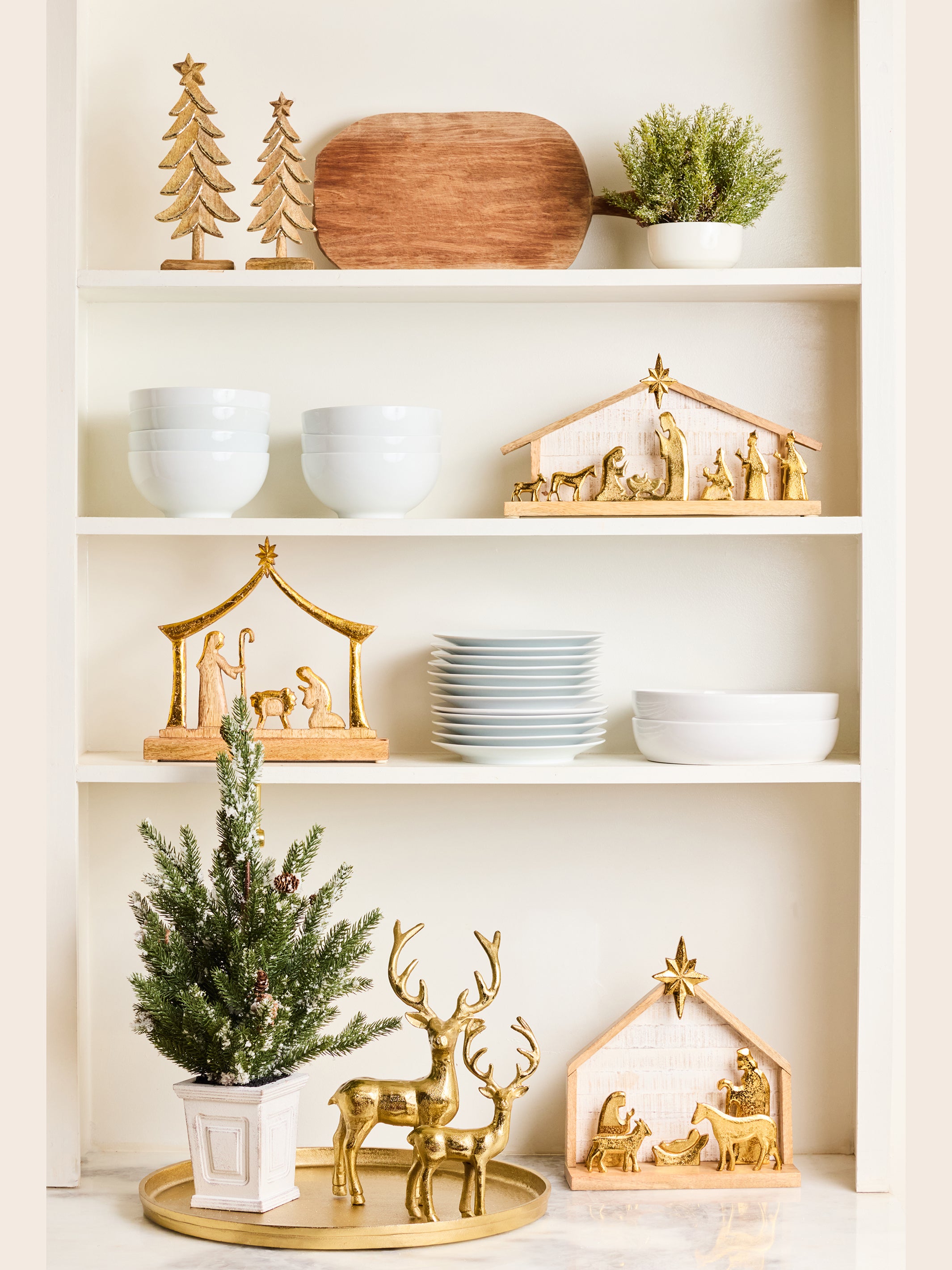 A styled holiday display on white shelves featuring gold nativity decorations, including a gold reindeer figurine, gold stable scene, and gold Christmas tree ornaments, alongside white ceramic bowls, plates, a small potted green plant, and a wooden cutting board.