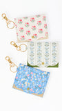 Three small fabric card holders or wallet keychains with gold-tone clasp rings, arranged vertically on a white background. The top one is cream with pink floral print, the middle one is white with blue floral print and green trim, and the bottom one is light blue with pink and white floral print.