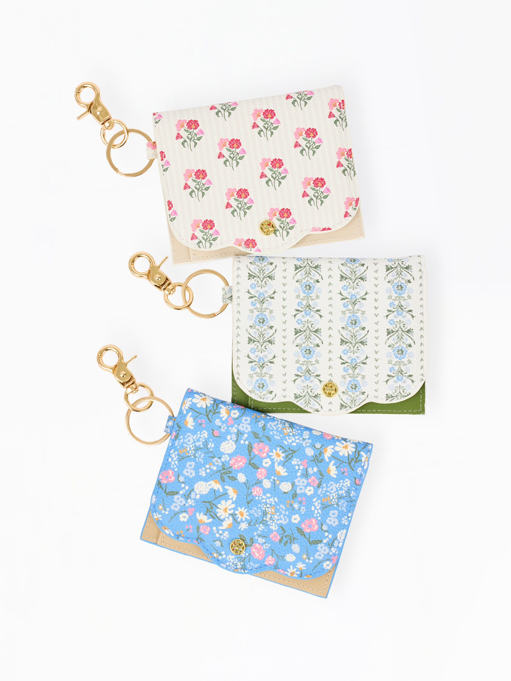 Three small fabric card holders or wallet keychains with gold-tone clasp rings, arranged vertically on a white background. The top one is cream with pink floral print, the middle one is white with blue floral print and green trim, and the bottom one is light blue with pink and white floral print.