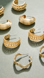 A styled flat-lay of multiple gold-toned hoop and cuff earrings arranged on a matte gray-green surface. The earrings feature various textures, including ribbed bands, woven patterns, and wavy open hoops, with some adorned with small clear stones.