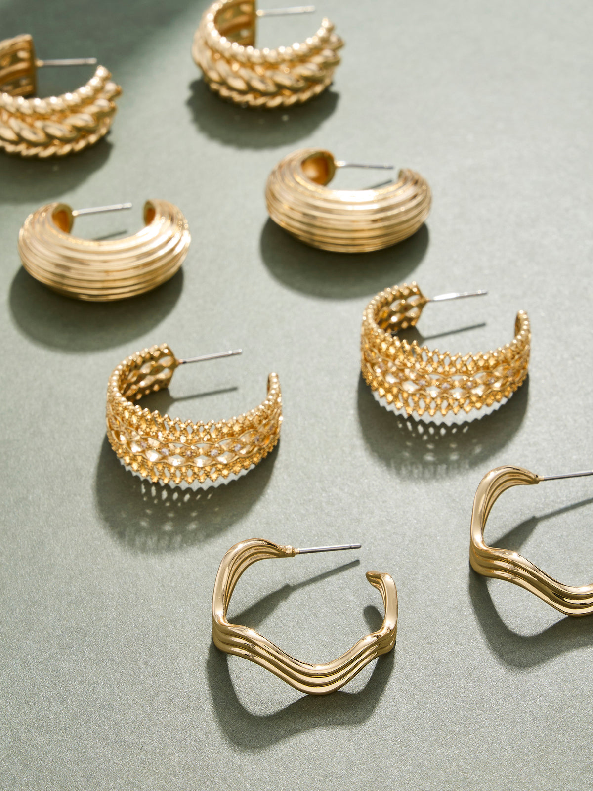 A styled flat-lay of multiple gold-toned hoop and cuff earrings arranged on a matte gray-green surface. The earrings feature various textures, including ribbed bands, woven patterns, and wavy open hoops, with some adorned with small clear stones.