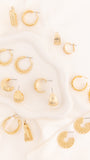 A flat-lay arrangement of multiple gold-toned hoop, crescent, and tassel earrings on a white draped fabric background.