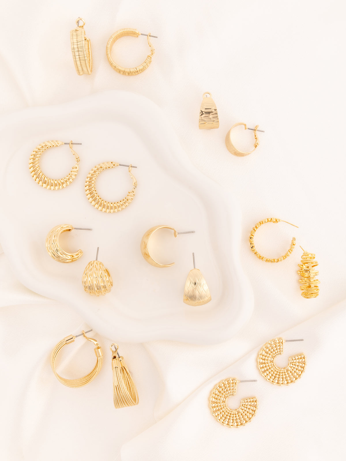 A flat-lay arrangement of multiple gold-toned hoop, crescent, and tassel earrings on a white draped fabric background.