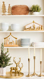 A styled holiday shelf display featuring gold nativity sets, white ceramic bowls and plates, a wooden cutting board, a small potted green plant, gold deer figurines, and three gold candlesticks against a white shelving unit.