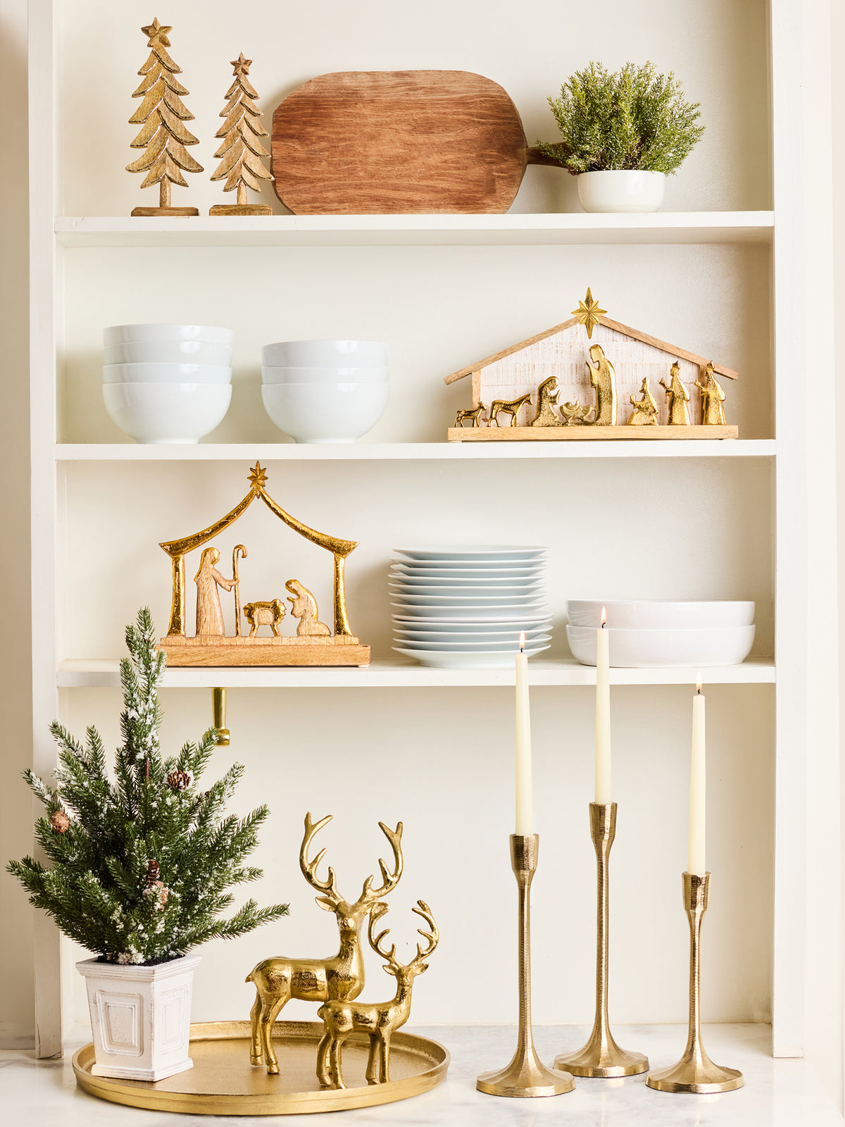 A styled holiday shelf display featuring gold nativity sets, white ceramic bowls and plates, a wooden cutting board, a small potted green plant, gold deer figurines, and three gold candlesticks against a white shelving unit.