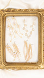 A gold ornate picture frame with a clear glass front, displaying several gold and crystal drop earrings and chains, set against a draped white satin background.