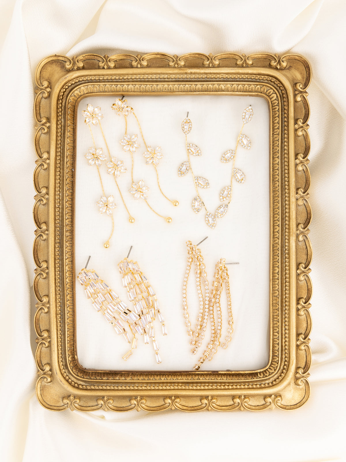 A gold ornate picture frame with a clear glass front, displaying several gold and crystal drop earrings and chains, set against a draped white satin background.